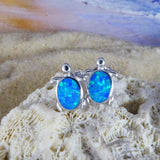 The Jewelry Network Caribbean Opal Turtle Stud Earrings feature silver turtle shapes with bright blue opal-like stones and a rhodium plated finish, shown on coral against a blurred sandy beach and ocean backdrop.