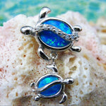 The Jewelry Network Caribbean Opal Double Turtle/CZ Line-Pendant features two silver turtles with bright blue synthetic opal shells on a textured pale coral background and a soft blue-green backdrop.