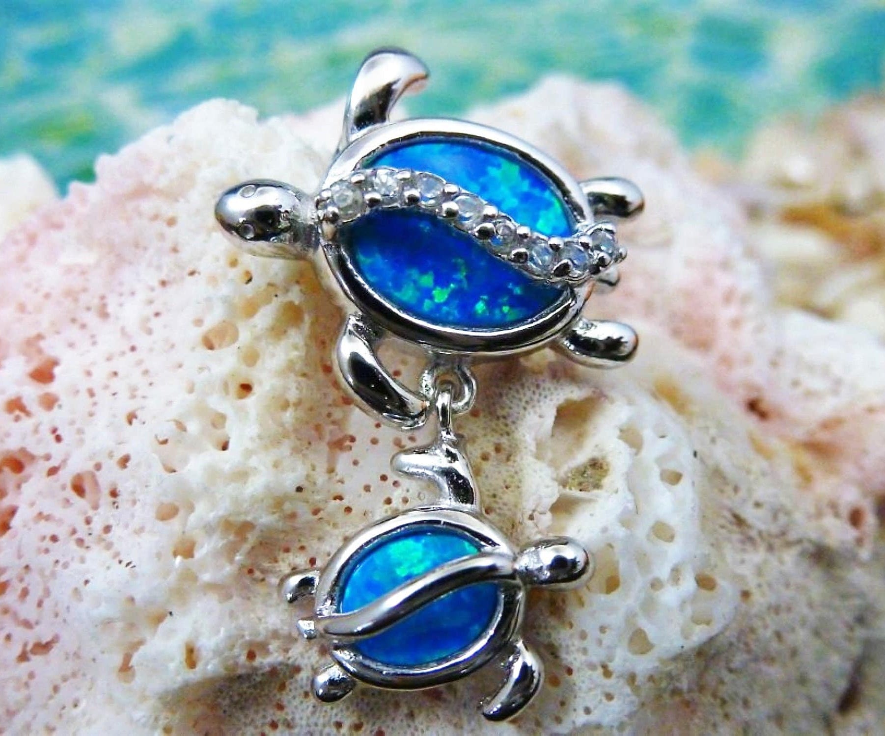 The Jewelry Network Caribbean Opal Double Turtle/CZ Line-Pendant features two silver turtles with bright blue synthetic opal shells on a textured pale coral background and a soft blue-green backdrop.
