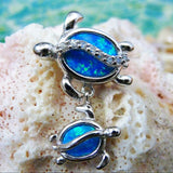 The Jewelry Network Caribbean Opal Double Turtle/CZ Line-Pendant features two silver turtles with bright blue synthetic opal shells on a textured pale coral background and a soft blue-green backdrop.
