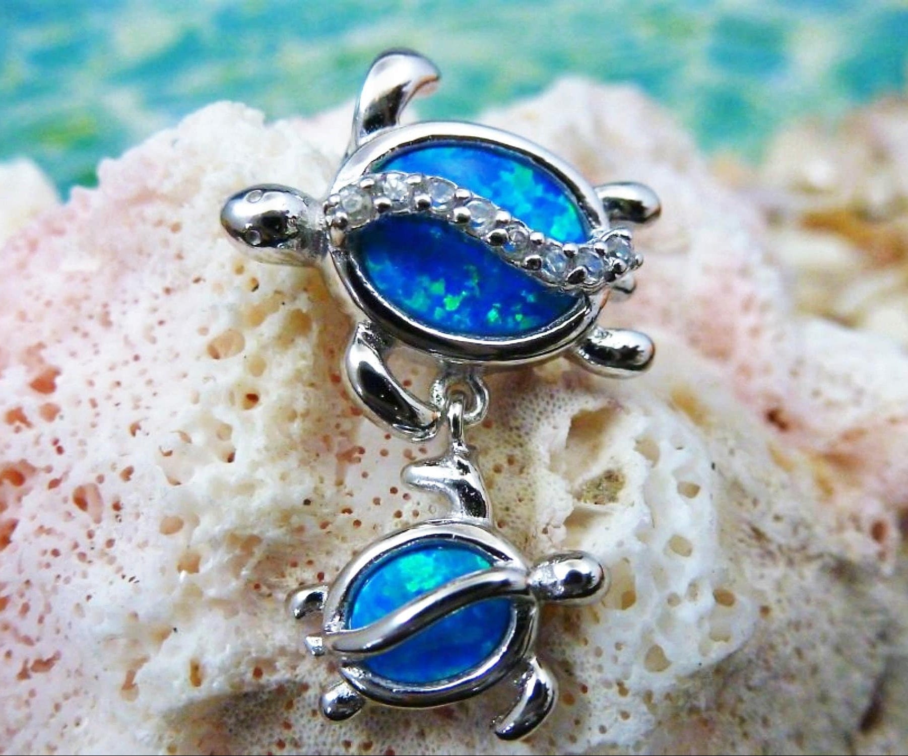 The Jewelry Network Caribbean Opal Double Turtle/CZ Line-Pendant features two silver turtles with bright blue synthetic opal shells on a textured pale coral background and a soft blue-green backdrop.