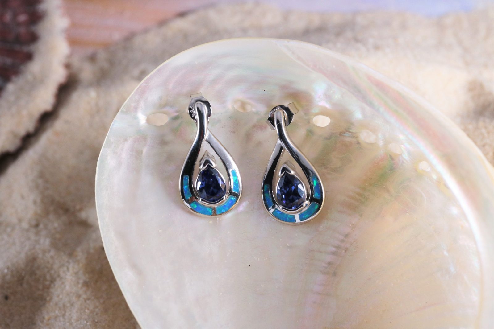 The Jewelry Network Caribbean Opal 4 Segment Teardrop/Tanzanite CZ Teardrop Post Earrings rest on a shiny, iridescent seashell, accented by sand.