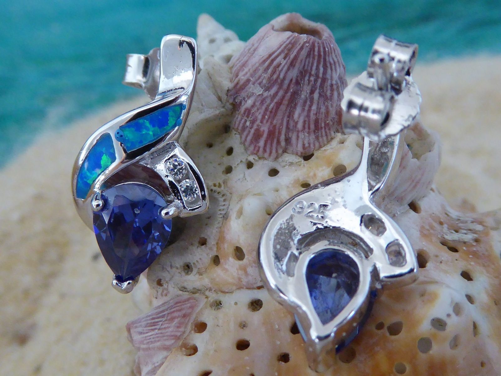 The Caribbean Opal 2 Segment Swish/CZ/Pear Amethyst CZ Earrings by Jewelry Network are shown on a textured seashell, set against a soft-focus sandy background with turquoise water accents.