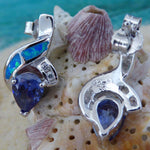 The Caribbean Opal 2 Segment Swish/CZ/Pear Amethyst CZ Earrings by Jewelry Network are shown on a textured seashell, set against a soft-focus sandy background with turquoise water accents.