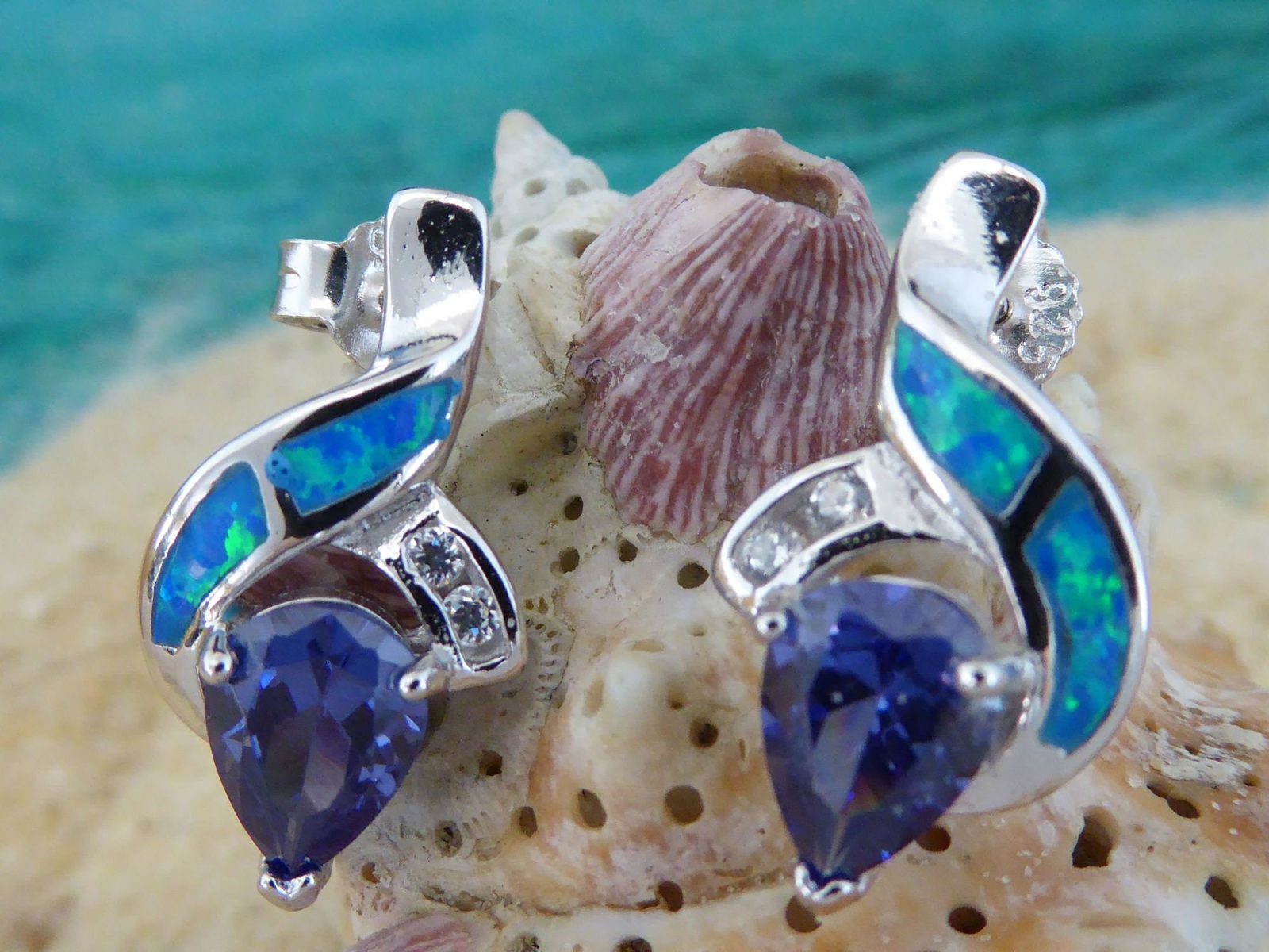 Jewelry Network’s Caribbean Opal 2 Segment Swish/CZ/Pear Amethyst CZ Earrings, featuring blue and green opal inlays with teardrop-shaped purple amethyst CZ stones, are shown against a seashell with a blurred ocean backdrop.