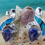Jewelry Network’s Caribbean Opal 2 Segment Swish/CZ/Pear Amethyst CZ Earrings, featuring blue and green opal inlays with teardrop-shaped purple amethyst CZ stones, are shown against a seashell with a blurred ocean backdrop.