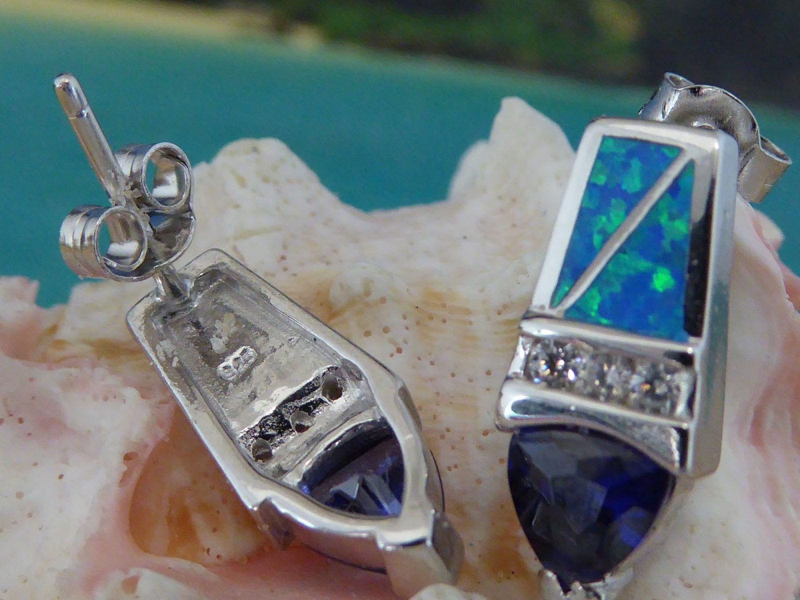 Jewelry Network's Caribbean Opal/CZ/Amethyst Point Earrings feature blue and green opal inlays and faceted blue gemstones, displayed on a light pink seashell with a blurred beach and ocean background.