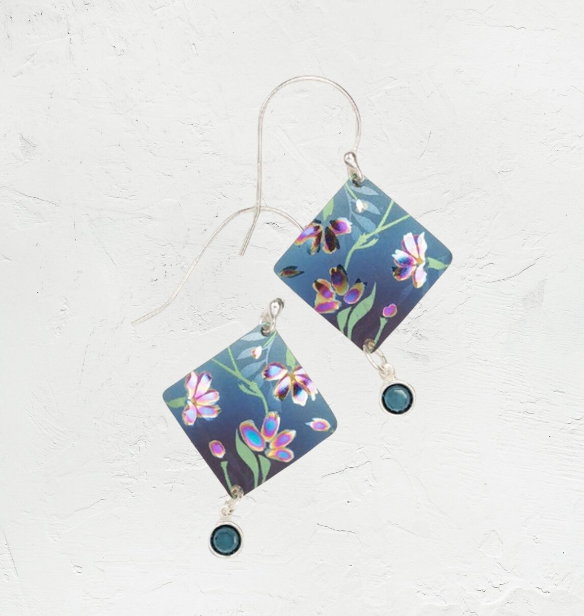 The Holly Yashi Garden Sonnet Blue Earrings showcase floral charm with square-shaped, nature-inspired pieces in blue and purple hues, accented by a round teal gemstone resting elegantly against a textured off-white background.