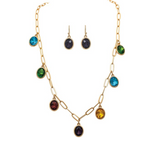 A-Train’s Gold Chain-link Multicolor Gem Drops Necklace Set features a vibrant gold chain necklace adorned with colorful oval gemstone pendants in green, blue, red, purple, and yellow. The set is perfectly paired with matching purple stone dangle earrings and is elegantly displayed on a white background.