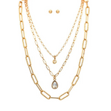 Displayed against a white background, the A-Train Gold Three Layered Chain Crystal Drops Necklace Set features three gold chain necklaces of varying lengths—each with a unique pendant, including a striking crystal drops design—paired with small gold stud earrings for an elegant layered look.