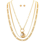 The A-Train Gold Multi Metal Chain Layer with Charms Necklace Set showcases four unique chain styles, including a pendant accented with a pearl and gold charm. Presented on a white background alongside coordinating gold stud earrings, this sophisticated jewelry set brings versatile elegance to any outfit.