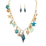 Introducing the A-Train Gold She Sells Seashells Charm Necklace Set—an elegant collection featuring ocean-inspired gold charms like shells, a crab, starfish, pearls, and blue beads. The set includes matching blue seashell-shaped dangle earrings, all beautifully displayed on a white background.