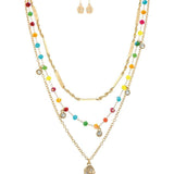 The A-Train Gold Multi-Color Fancy Chain Layer Necklace Set includes layered gold necklaces with colorful beads and sparkling pendants, paired with matching earrings featuring small gold skull charms.