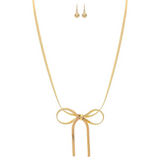 The A-Train Gold Herringbone Chain Bow Tie Necklace Set features a central ribbon bow design, matching gold stud earrings, and is crafted with mixed jeweler’s metals, shown on a white background.