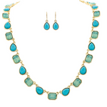 The A-Train Gold Blue Glow Gem Shapes Necklace Set features a necklace with alternating blue teardrop and rectangular stones and matching teardrop earrings, elegantly displayed on a crisp white background.