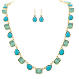The A-Train Gold Blue Glow Gem Shapes Necklace Set features a necklace with alternating blue teardrop and rectangular stones and matching teardrop earrings, elegantly displayed on a crisp white background.