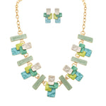 The A-Train Gold Multi-Color Green Layered Blocks Necklace Set features overlapping rectangular pieces in green, blue, yellow, and pearl white for a bold look, with matching earrings included.