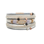 The A-Train Gold White Druzy Cord Magnetic Bracelet pairs gray cord, small white beads, black and rose gold accents, and a central oval opalescent stone for a stylish, multi-layered look.