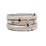 The A-Train Gold White Druzy Cord Magnetic Bracelet pairs gray cord, small white beads, black and rose gold accents, and a central oval opalescent stone for a stylish, multi-layered look.