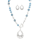 The A-Train Silver Blue Frosty Bead Teardrop Topple Necklace Set features a silver and blue beaded necklace with a large silver teardrop pendant and matching textured oval earrings, displayed on a white background.