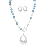 The A-Train Silver Blue Frosty Bead Teardrop Topple Necklace Set features a silver and blue beaded necklace with a large silver teardrop pendant and matching textured oval earrings, displayed on a white background.