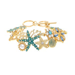 The A-Train Gold Enamel Crystal Seaside Mosaic Toggle Clasp Bracelet features sea-inspired charms—seashell, starfish, pearl, blue gemstones—set on white enamel and adorned with sparkling crystals.
