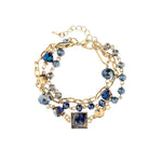 A-Train’s Gold Glass & Acrylic Squares Link Bracelet features multiple gold chains with blue and iridescent beads, gold charms, and a marbled blue-and-white square centerpiece. An adjustable clasp ensures a perfect fit.