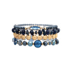 The A-Train Gold Dark Blue Stone Bead Glass 4 Pc Set Bracelet features stylish dark blue, gold, and light blue beads with a central oval gold dark blue stone accent, shown on a white background.