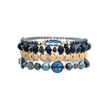 The A-Train Gold Dark Blue Stone Bead Glass 4 Pc Set Bracelet features stylish dark blue, gold, and light blue beads with a central oval gold dark blue stone accent, shown on a white background.