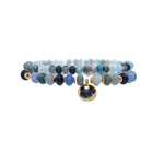 The A-Train Gold Blue Color Treated Jade Bead 2 Pc Set Bracelet features shades of blue and teal beads, gold accents, and a round gold charm with a blue stone at the center.