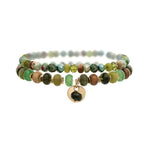 The A-Train Gold Green Color Treated Jade Bead 2 Pc Set Bracelet features two strands of multicolored beads and a gold charm with a dark stone, perfect for everyday or special occasions. Shown on a white background.
