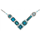 The La Dolce Vita “V” Moonlight Light Blue Necklace by Firefly features a silver V-shaped pendant adorned with circular and square turquoise and blue enamel designs, accented with small blue gems. This stunning handmade piece of jewelry adds an elegant touch to any ensemble.