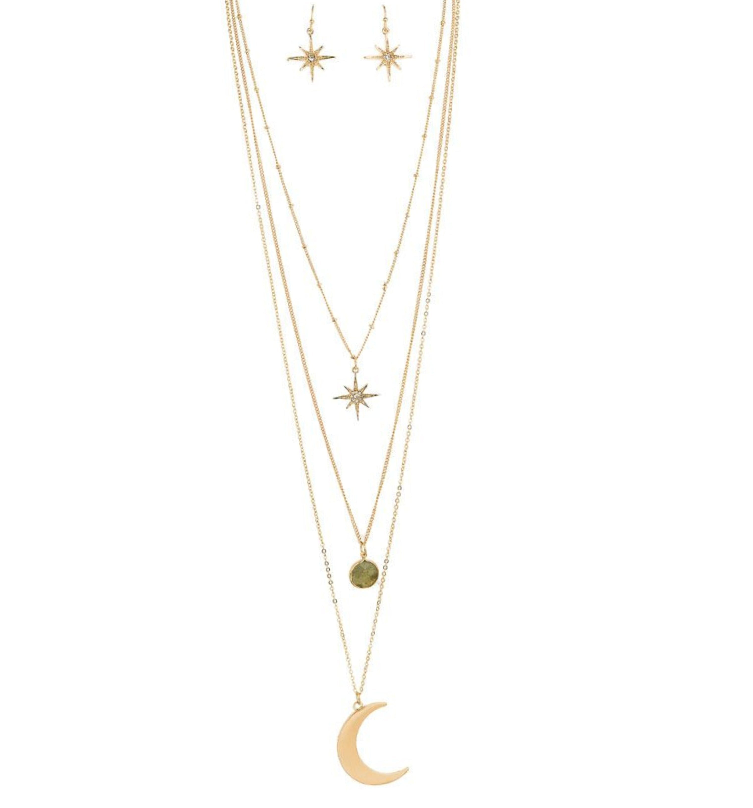 The A-Train Gold Three Layer Moon/Star Necklace Set features star-shaped earrings and a layered necklace with starburst, green stone, and crescent moon pendants for a chic, versatile look.