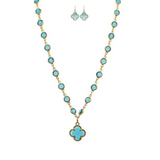 A-Trains Gold Blue Crystal By The Inch Clover Charm Necklace Set features a gold necklace with turquoise stones and a clover charm, paired with matching turquoise and gold dangle earrings, beautifully displayed on a white background.
