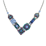 The La Dolce Vita “V” Light Blue Necklace by Firefly features a silver V-shaped pendant with blue, purple, and teal gemstones in ornate designs—a handcrafted statement piece from Guatemala.
