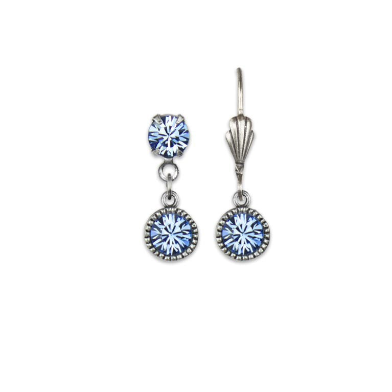 The Simple Drop Light Sapphire Earrings by Anne Koplik Designs, Inc. are silver-toned with a timeless look—one earring has two round light blue stones, the other features a single gemstone and a decorative silver accent at the top.