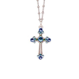 The Fancy Stone Cross Blue Necklace by Anne Koplik Designs, Inc. is a silver plated necklace with a budded cross pendant featuring blue and green crystal accents, shown on a white background.